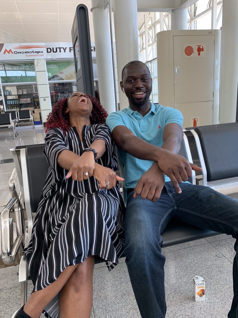 Kemi and Pelu sitting at an airport and laughing while crossing their hands (and showing off an engagement ring)